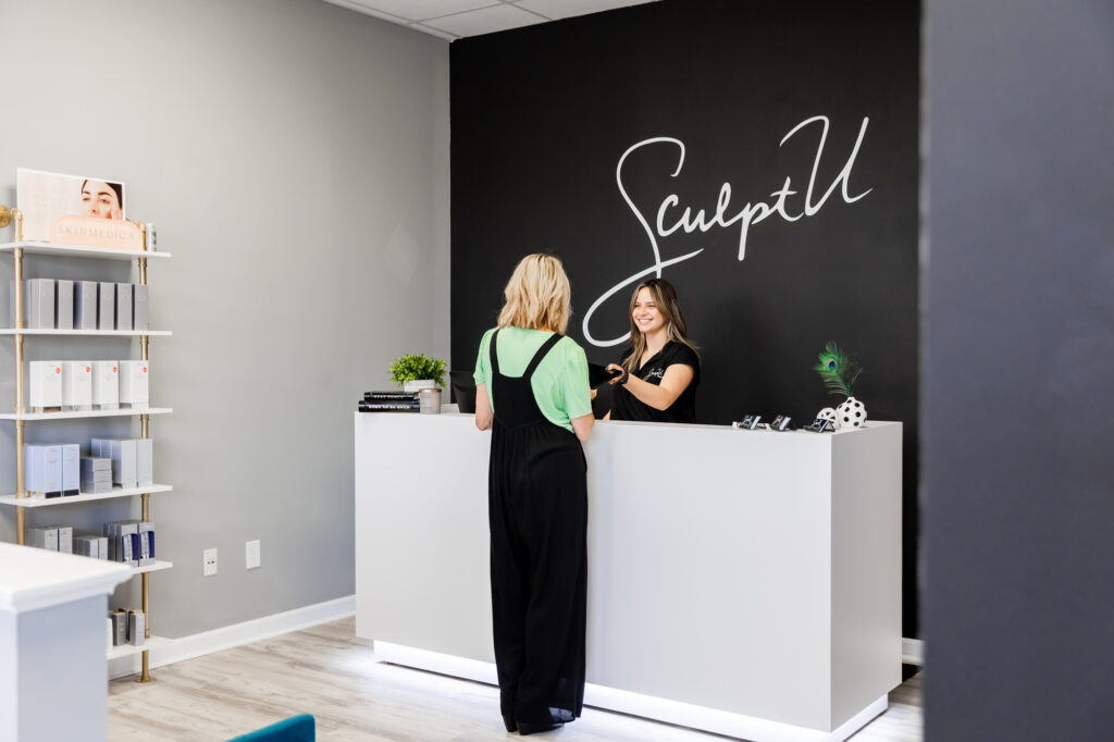 A wide shot of a patient checking into her eye treatment in Louisville appointment with the friendly front-desk associate at SculptU. The associate hands the patient some pre-appointment paperwork to fill out, smiling as she does.